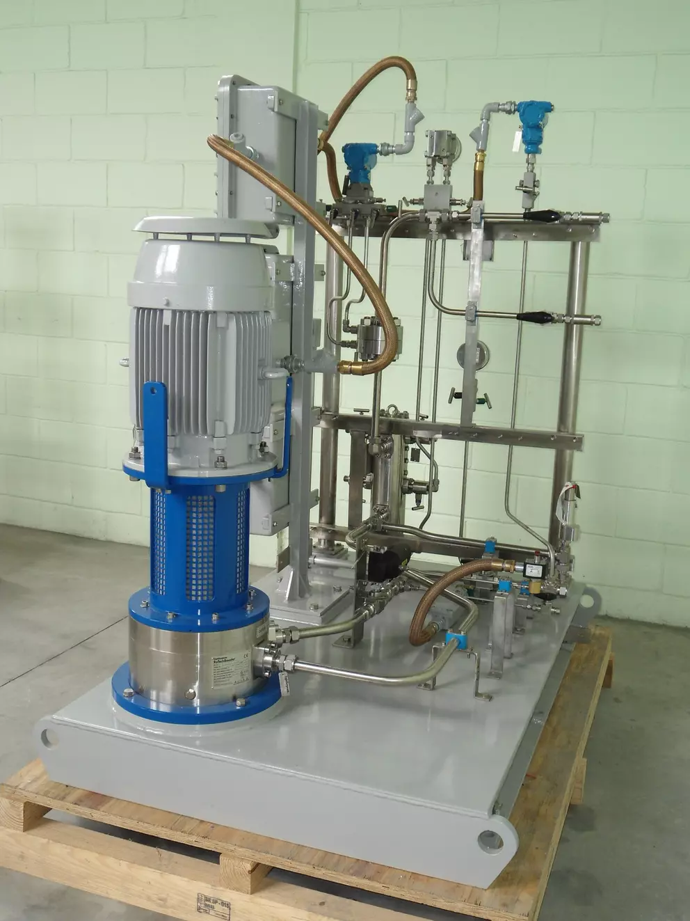 Nahaufnahme einer Industriemaschine, wahrscheinlich einer Schmieröl- oder Hydraulikaggregat, mit einem großen grau-blauen Elektromotor und einer Pumpenbaugruppe, die an eine Edelstahlrohrleitung und ein Ventilgestell angeschlossen sind.