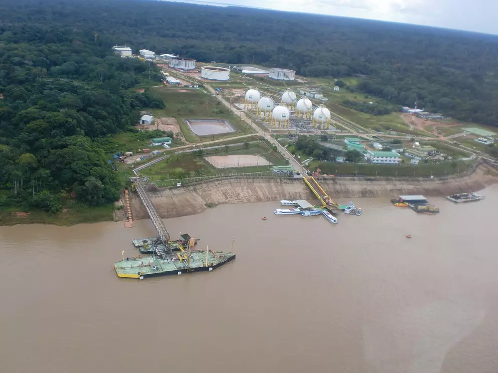Luftaufnahme einer Öl- und Gasförderanlage an einem Fluss mit Lagertanks, Gebäuden und Booten, umgeben von dichtem Regenwald.